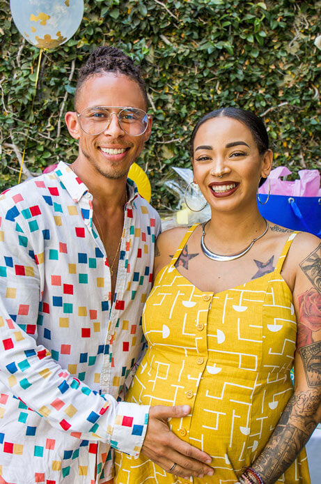 A happy couple stands in front of a hedge. A pregnant person's partner holds their belly