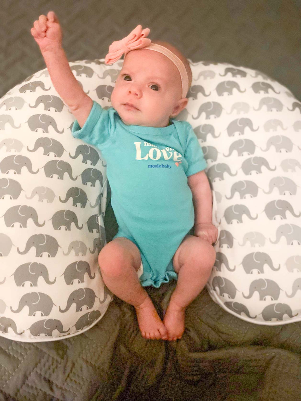 6 week old baby girl in a blue Mosie Baby onesie raises on fist in the air and lays on a baby pillow with gray elephants. She has a pink bow and headband on her head.