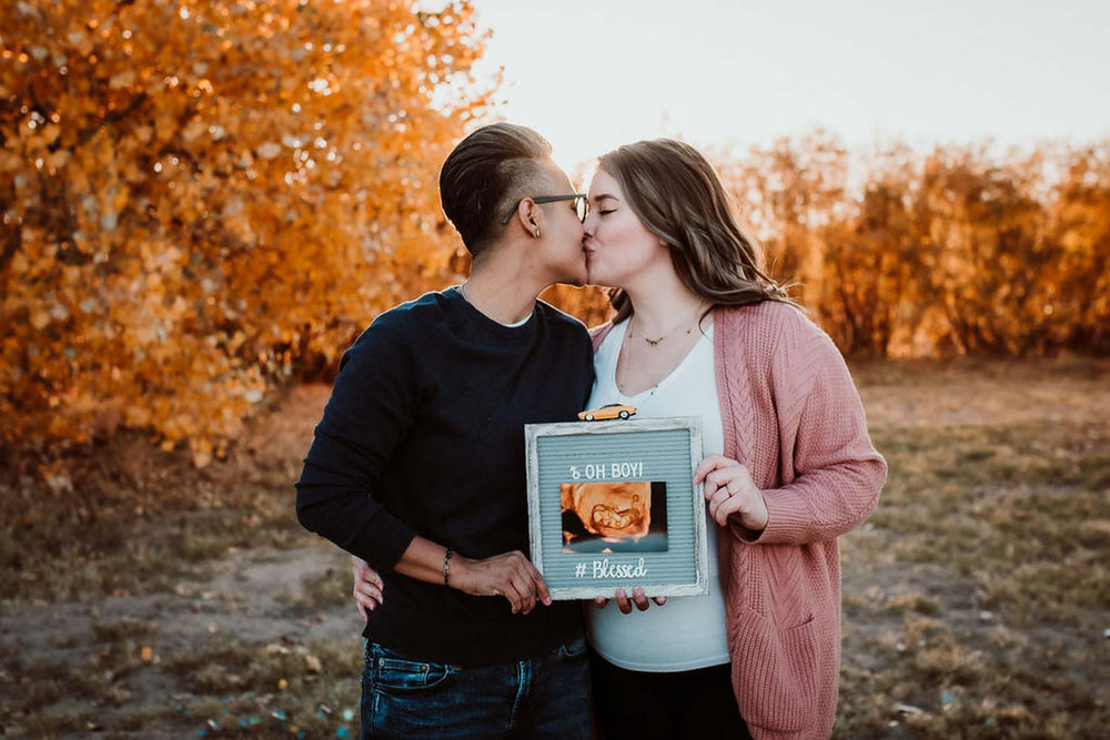 Two women kiss in front of fall trees while holding their baby announcement sign reading "Oh Boy" with a sonogram of their baby.