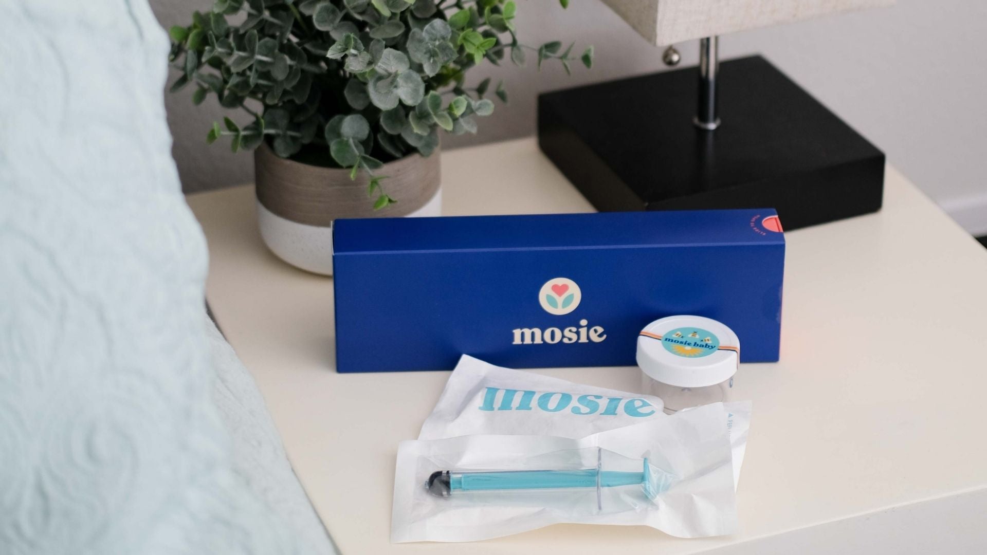 The Mosie Kit opened with both Mosie syringes and collection cup on a bedside table with plant and lamp